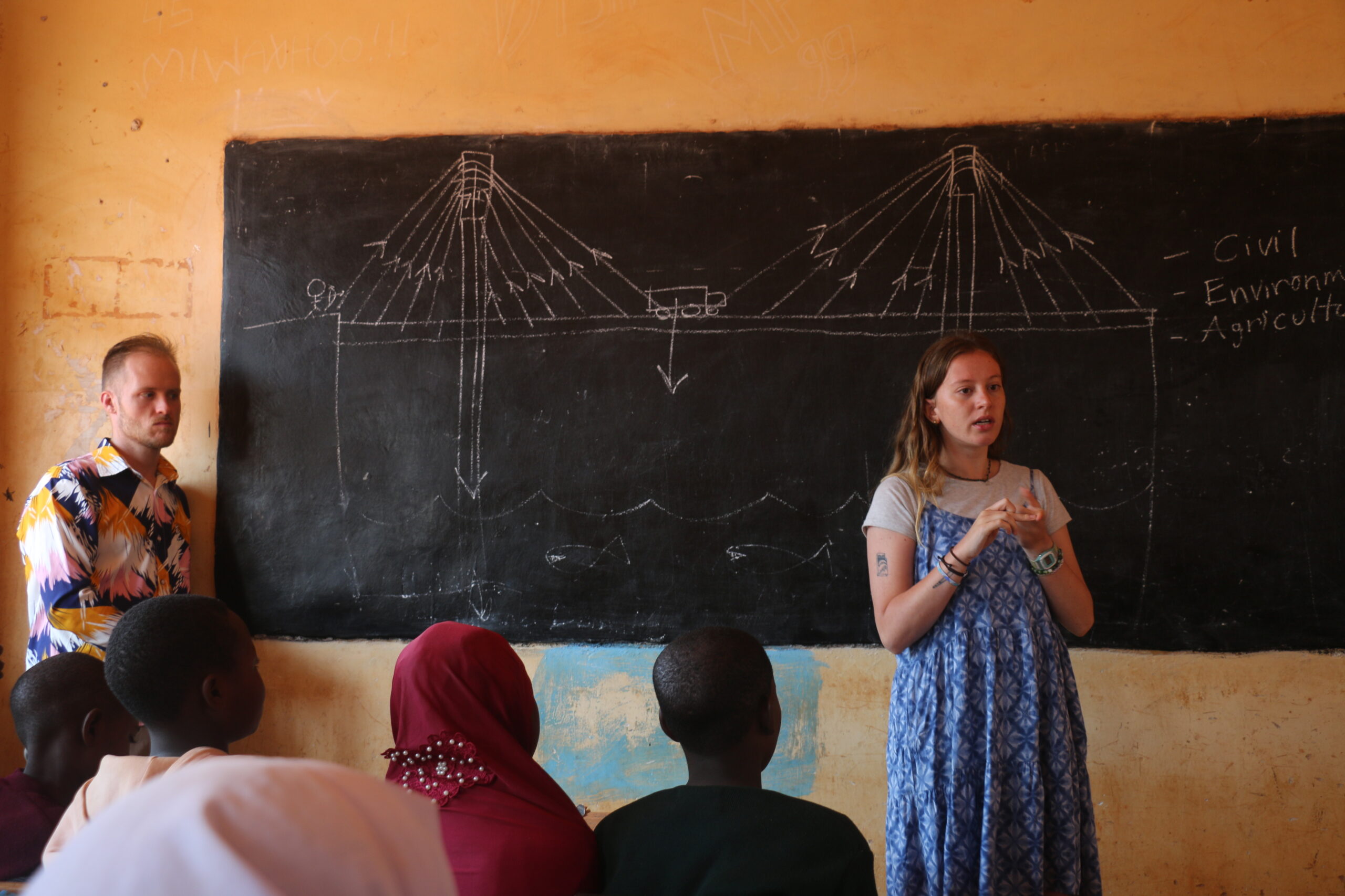 A woman and a man stand in front of a chalkboard in a classroom with students facing them.