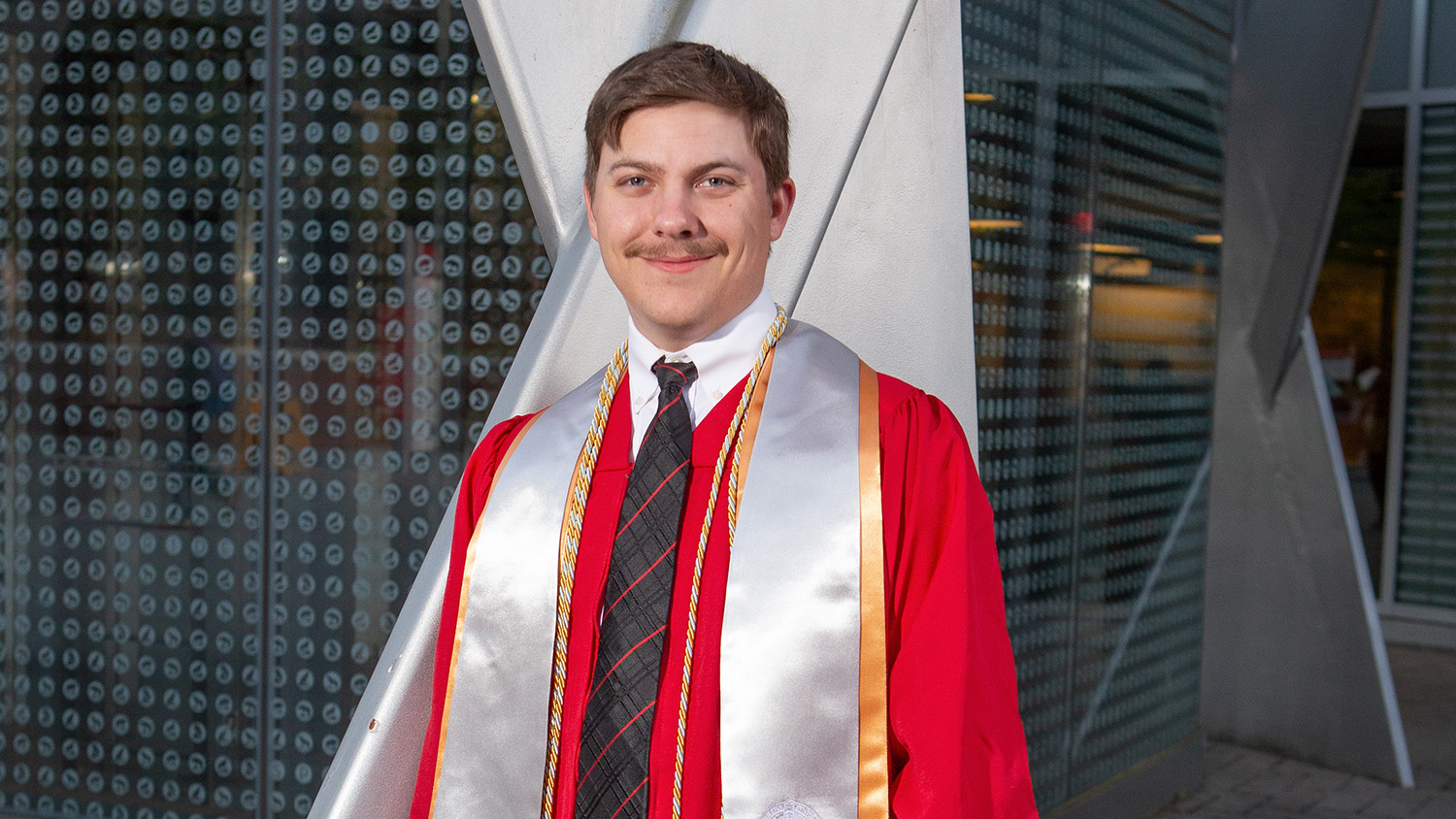 a man wearing a red graduation gown with yellow and gray cords and a black and red striped tie