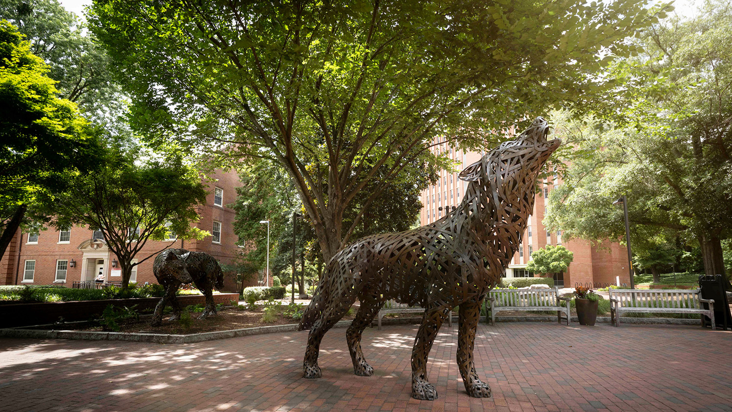 two wolf statues made of copper weaved metal standing on a brick patio surrounded by trees
